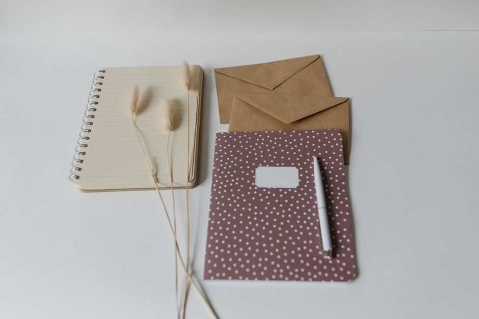 Artistic flat lay featuring notebooks, brown envelopes, and decorative bunny tail grass on a white background.