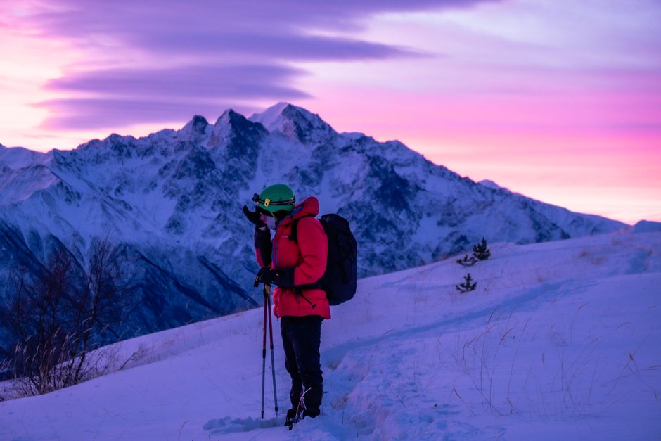 Person hiking in winter gear on a snowy mountain with a vibrant sunrise backdrop.