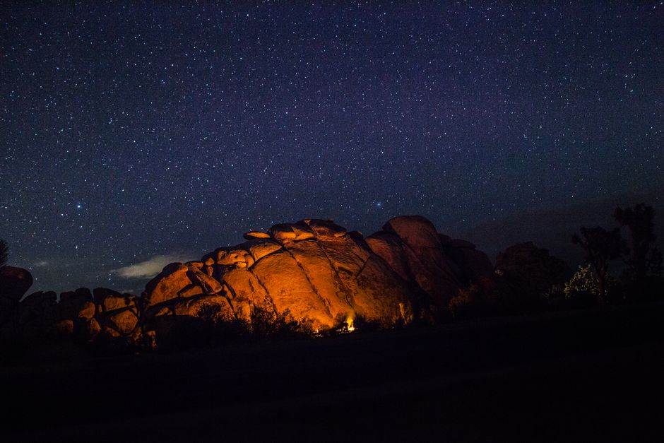 A campfire casts a warm glow on rocky formations under a star-filled night sky.